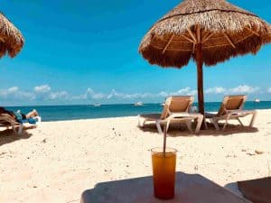 An orange cocktail and empty chairs on the beach under the shadow on the white sand by the water of crystal clear sea during sunny warm day; best beach clubs in malaga