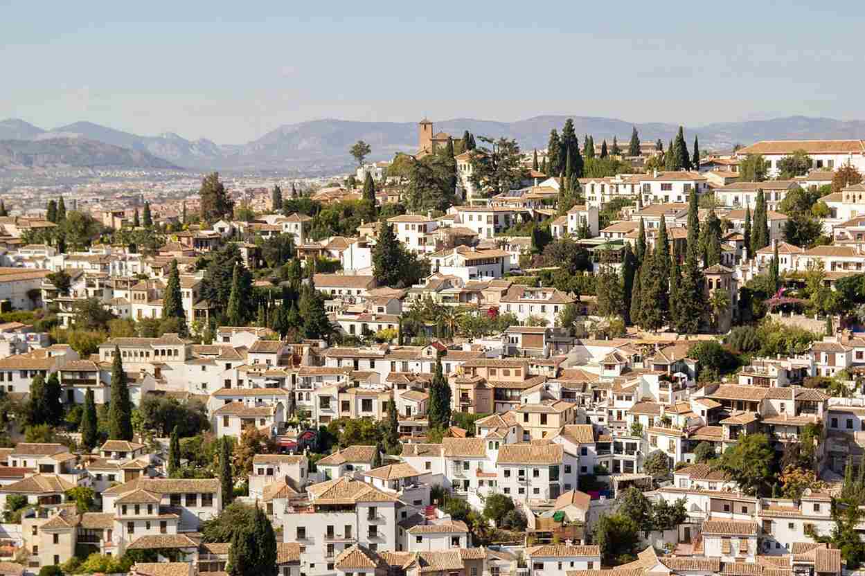 The view from above on various white houses and buildings of Albaicin, the neighborhood in Granada, Spain and some green trees between the houses during the sunny day.