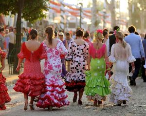 Five young women dressed in colorful Flamenco dresses during Feria Malaga. Random crowd of people in the blurry background during sunny summer day.