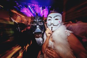 Halloween party in the club, people dressed as different characters wearing some masks on the blurry background with purple lights.