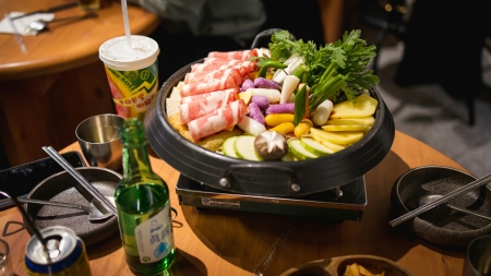 A Korean hotpot dish called budae jjigae containing various raw meat and vegetables simmering on an electric burner on a dining table.