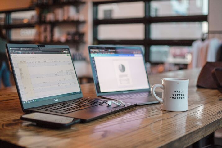 Two laptops and a phone are on a table in a coworking space, with a cup of coffee beside them.