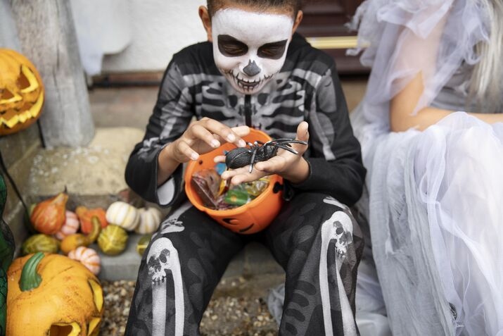 A child in a skeleton costume strokes a plastic spider while a can of Halloween candies rests on their legs.
